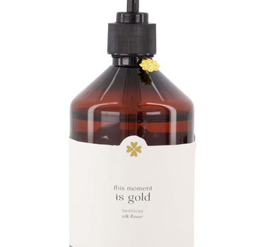 "This Moment Is Gold" hand soap by Zusss in a white bottle with an amber-colored body, a black pump, and a gold floral accent